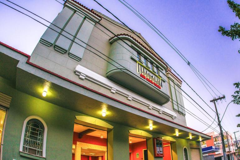 Fotografia em ângulo baixo da fachada do Cine Santa Tereza, um prédio histórico. No andar superior, há uma varanda curva sob pequenas janelas com o letreiro dourado "CINE SANTA TEREZA" iluminado. Luzes quentes embutidas na marquise iluminam a calçada, enquanto cabos elétricos cruzam o céu azul ao fundo.