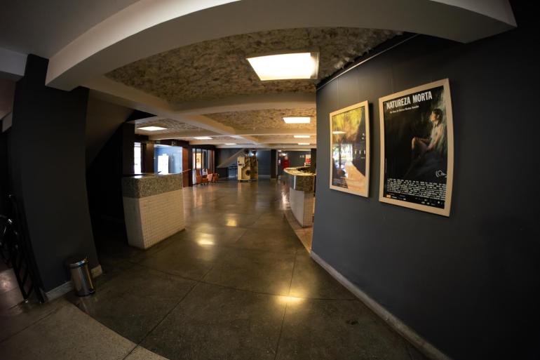 CineBH e Brasil CineMundi debatem políticas públicas para o audiovisual da capital Hall de entrada amplo e iluminado do Cine Santa Tereza, com piso cinza reluzente, luminárias quadradas no teto e cartazes de filmes pendurados em uma parede cinza-escuro à direita, incluindo o do filme "Natureza Morta".