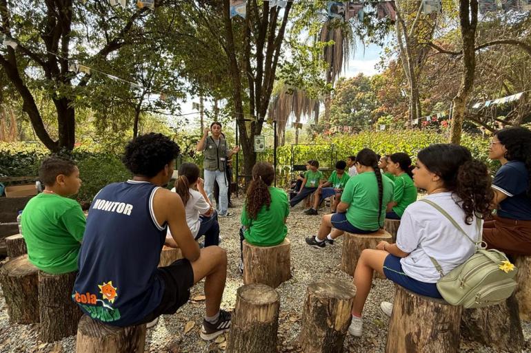 Imagem de um grupo de crianças sentadas recebendo informações sobre meio ambiente, em espaço aberto, embaixo de uma árvore