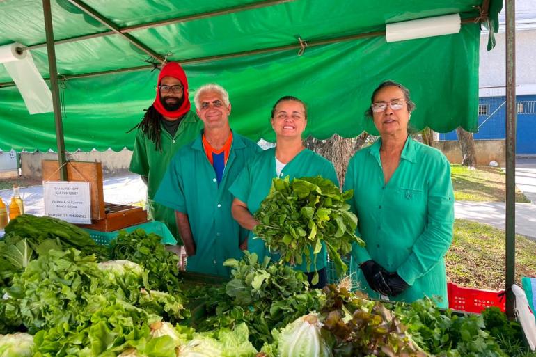 PBH celebra os profissionais que movimentam a economia na cidade no Dia do Feirante Quatro feirantes postados na banca, usando roupa verde, com as verduras expostas no primeiro plano.