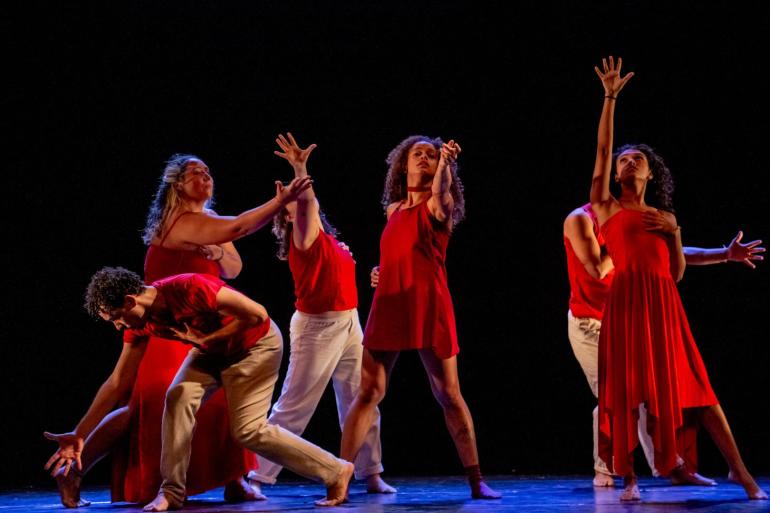 Cena do espetáculo de dança "Sangue Latino" em um palco escuro. Seis dançarinos descalços vestem figurinos em tom vermelho e branco. Com expressões dramáticas e mãos abertas, eles realizam movimentos expansivos: alguns em pé com os braços erguidos para o alto, e um homem agachado à esquerda inclinado sobre o chão.