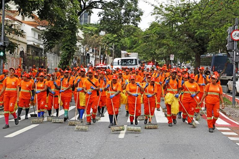 SLU celebra 53 anos com exposição fotográfica e de equipamentos no Parque Municipal Imagem mostra grupo de garis vestidos com uniformes da SLU varrendo uma rua.