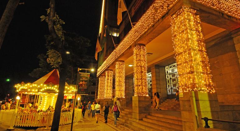 Fachada da Prefeitura Municipal de Belo Horizonte, com luzes natalinas e carrossel à frente, também iluminado, no início da noite. 