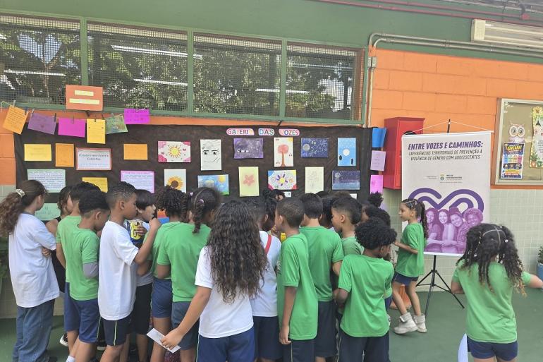 PBH promove ação comunitária de prevenção e enfrentamento da violência contra crianças e adolescentes Grupo de crianças de uniforme escolar reunidas de costas, a observar uma exposição de desenhos e cartazes na parede sob o título "Galeria das Vozes".