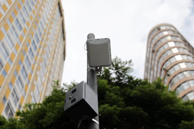 Praças e parques da capital ganham novos pontos do Wi-Fi BH Gratuito Imagem tirada de baixo para cima, pegando o alto de um poste com estrutura de wi-fi, tendo no fundo parte do prédio do antigo Hotel Othon, a copa de uma árvore e outro edifício da região central.