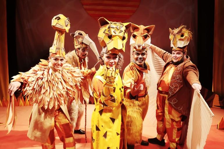 Peça no Teatro Marília reúne animais do Cerrado em defesa do bioma na imagem possuem cinco atores caracterizados como animais do bioma Cerrado. Vestindo figurinos em tons de amarelo, laranja e marrom, o elenco destaca-se pelas máscaras no estilo geométricas dispostas sobre a cabeça, representando diversos animais posicionados à frente de um painel ilustrado com o sol.