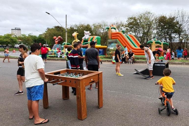 Imagem mostra uma rua com pula pula, totó, crianças brincando, entre elas um menino pedalando vestido com uma camiseta amarela.