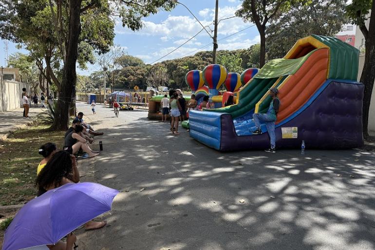 A Rua é Nossa recebe projetos especiais e promove atividades de lazer em seis regiões de BH Em uma rua arborizada e fechada para o trânsito, acontece uma edição do programa "A Rua é Nossa". À direita, há grandes brinquedos infláveis coloridos, entre eles um escorregador nas cores verde, laranja, azul e roxo e estruturas em formato de balões. Crianças e adultos circulam e brincam pelo espaço. Algumas pessoas acompanham as atividades em pé, enquanto outras estão sentadas à sombra das árvores, na calçada à esquerda. Em primeiro plano, uma pessoa se protege do sol com uma sombrinha roxa. Ao fundo, ap