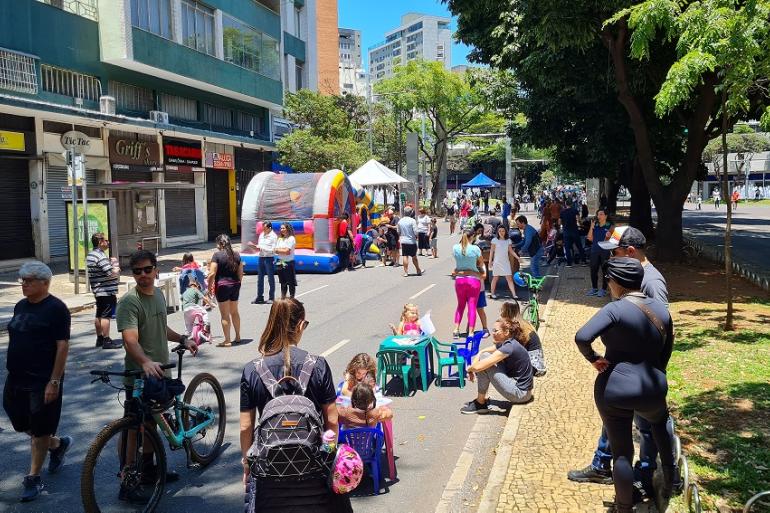 Programação do A "Rua é Nossa" deste domingo leva lazer, cultura e esporte para comunidades de Belo Horizonte Em uma rua fechada ao trânsito, durante um dia ensolarado, crianças e adultos participam de atividades de lazer. Ao centro, há brinquedos infláveis coloridos, tendas, pequenas mesas e cadeiras infantis. Algumas crianças brincam, enquanto outras pessoas caminham, conversam ou acompanham as atividades. Em primeiro plano, há pessoas com bicicletas e uma mulher com mochila próxima a um grupo de crianças. À direita, árvores grandes fazem sombra sobre a calçada, onde outros participantes estão reunidos. Ao fundo,