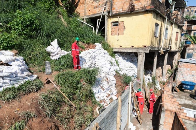 Imagem de servidores da Urbel trabalhando na construção de muro de contenção em área de encosta, com imóveis próximos