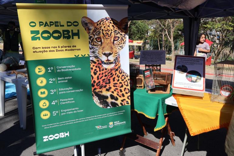 Imagem mostra um estande, onde há um banner alusivo ao tema: O papel do Zoo BH. Mostra também os cinco pilares dos zoos modernos: conservação, bem-estar animal, educação ambiental, pesquisa científica e lazer/recreação. Além do banner há também gaiolas e mesas. Uma criança aparece no fundo da foto.