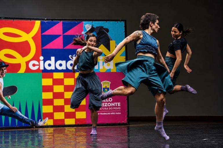 Oficinas de vogue, freestyle e circo são destaques da programação desta semana no Centro de Referência das Juventudes Fotografia horizontal de uma apresentação de dança em um palco com piso preto e brilhante. Três bailarinas executam movimentos coreografados. Em primeiro plano, à direita, uma bailarina de cabelos curtos, blusa e saia-calça azul-petróleo, com tatuagens nos braços, realiza um salto com uma das pernas estendida para trás. À esquerda, outra bailarina, de cabelos presos, veste blusa escura e calça larga, com um dos braços flexionado à frente do corpo. Ao fundo, à direita, a terceira bailarina, de blusa preta e