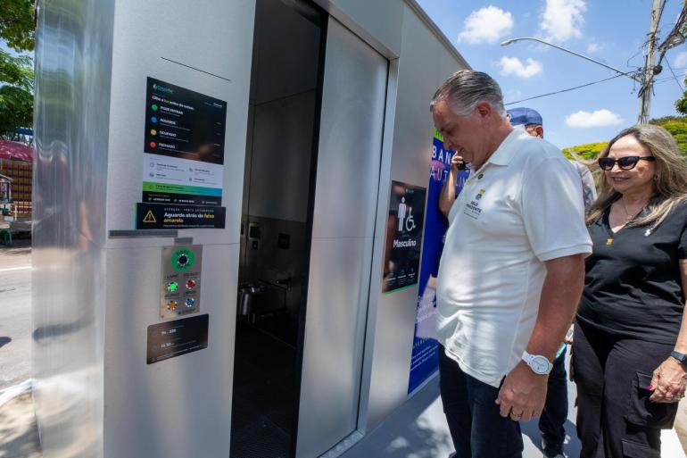 Prefeito, ao lado de uma mulher de preto, abre a porta do sétimo banheiro autolimpante da cidade.