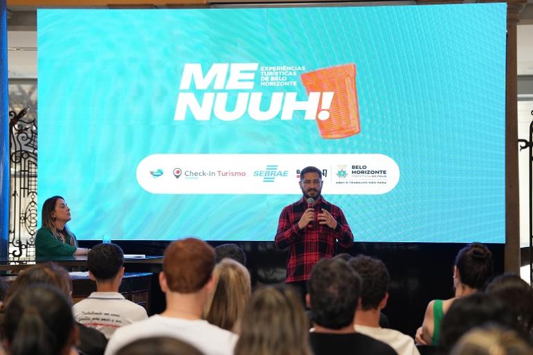 O presidente da Belotur, Eduardo Cruvinel, fala durante o lançamento da segunda etapa do "Menuuh! – Experiências Turísticas de Belo Horizonte". Ao fundo, o painel apresenta a identidade visual do projeto,