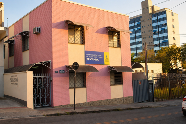 A imagem mostra a sede do Centro Especializado de Atendimento à Mulher. Trata-se de uma casa de dois andares com fachada predominantemente rosa, de tijolos aparentes. A parte inferior das paredes é revestida com pedras em tons de cinza. Na fachada há várias janelas protegidas por grades metálicas e cobertas por pequenas marquises curvas de metal. No centro, há uma placa azul e amarela com a identificação “Centro Especializado de Atendimento à Mulher Benvinda”, acompanhada da marca da Prefeitura de Belo Hor