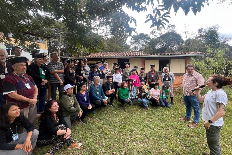 Agentes ambientais assistem aula em uma área verde. Em frente há uma mulher que gesticula e um homem postado ao lado dela.