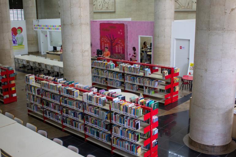 Estantes na horizontal, na cor vermelha, com vários livros coloridos. As estantes estão entre pilastras na cor marfim