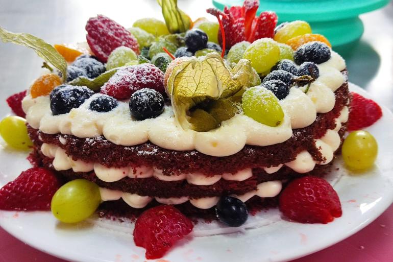 Inscrições abertas para 60 vagas em três cursos gratuitos do Trilhas da Gastronomia Imagem mostra um bolo Red Velvet inteiro, coberto por creme e frutas vermelhas e amarelas. O bolo está disposto em um prato branco.