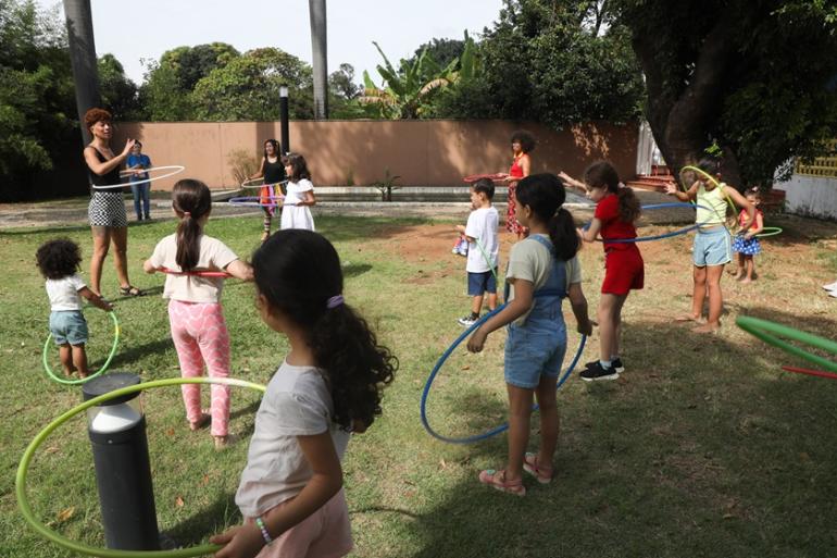Mês da Criança e Outubro Prateado movimentam a programação do “Museus Pampulha” A imagem mostra um grupo de pessoas, predominantemente crianças, participando de uma atividade de bambolê em um ambiente ao ar livre com gramado e vegetação ao fundo, no quintal do Museu Casa Kubitschek.