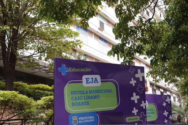 Escola Municipal Caio Líbano Soares celebra 35 anos transformando trajetórias na EJA Placa roxa em primeiro plano exibe o texto "Educação EJA Escola Municipal Caio Líbano Soares" junto ao brasão da Prefeitura de Belo Horizonte. Ao lado, há outra placa similar. Ao fundo, vê-se a fachada de um prédio claro de vários andares com janelas e detalhes em azul, cercado pelas folhagens e galhos verdes de árvores frondosas que emolduram a parte superior da imagem.