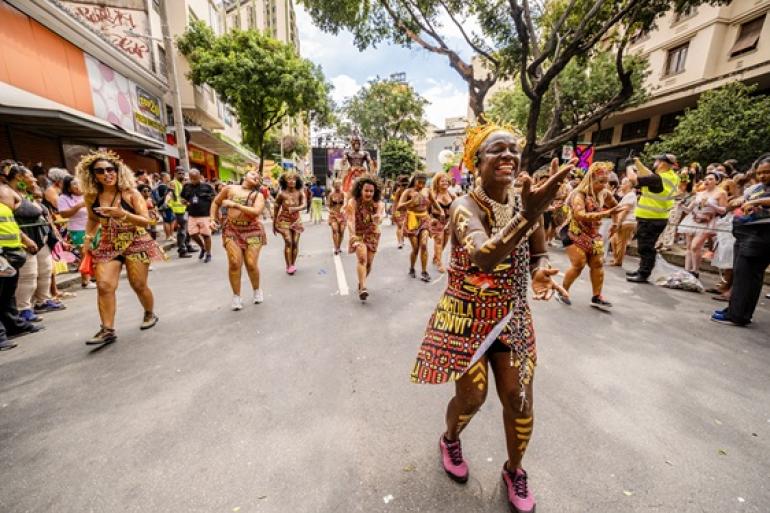 Integrantes do bloco Angola Janga desfilam durante o Carnaval de Belo Horizonte, com destaque para uma das integrantes à frente do cortejo, enquanto foliões acompanham a apresentação pelas ruas da cidade.