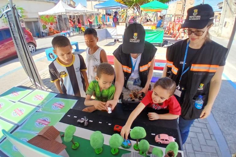 Mutirão de limpeza no Morro do Papagaio reúne a PBH, órgãos parceiros e a comunidade Foto de um evento comunitário ao ar livre, com quatro crianças participando de uma atividade em uma mesa, acompanhadas por duas servidoras uniformizadas. Sobre a mesa, há uma maquete com árvores, casas e veículos de brinquedo. Ao fundo, aparecem tendas, pessoas e materiais do evento.