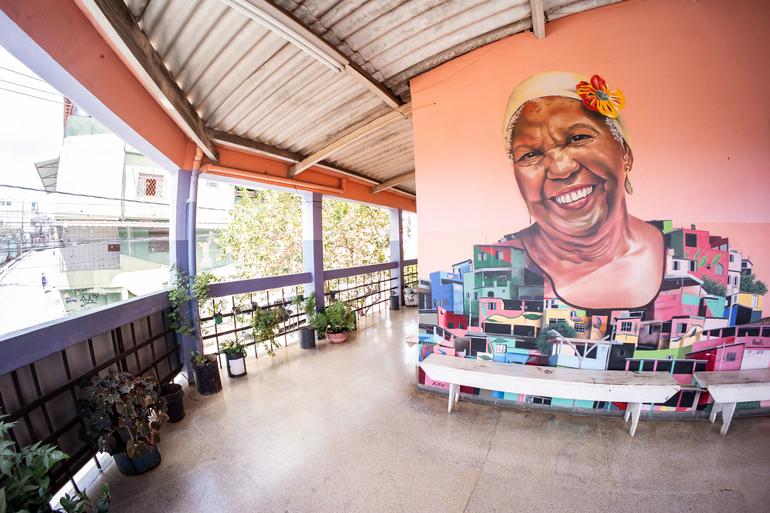 Mês de agosto terá atrações variadas nos centros culturais São Geraldo, Alto Vera Cruz e na Usina de Cultura Foto do Centro Cultural Alto Vera Cruz. A imagem mostra uma varanda coberta, com piso claro, vasos de plantas ao longo do guarda-corpo e bancos brancos. Na parede ao fundo, há um grande mural colorido com o retrato de uma mulher negra sorridente, usando um lenço na cabeça com uma flor. A pintura também retrata casas em tons vibrantes, remetendo à paisagem da comunidade do Alto Vera Cruz. Ao lado da varanda, é possível ver parte da rua e das construções do entorno.
