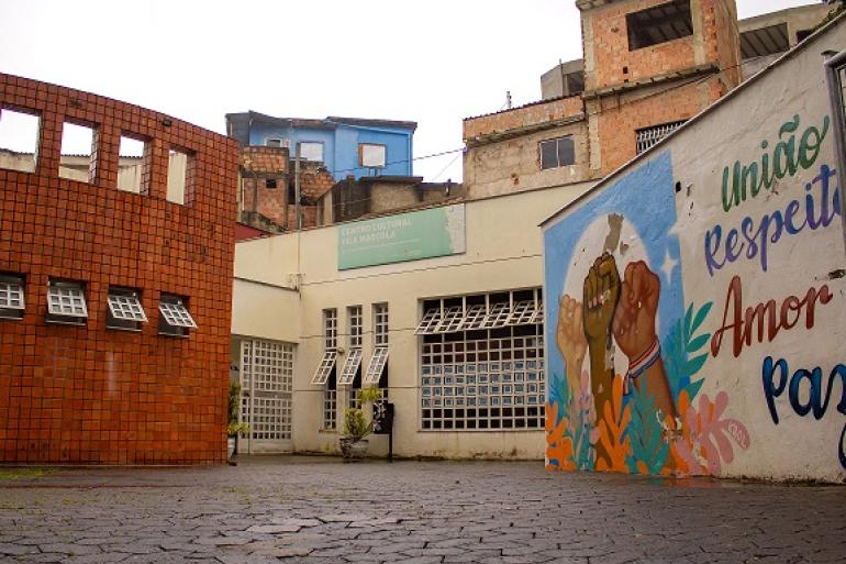 Centros Culturais Vila Fátima e Vila Marçola celebram o patrimônio cultural com atividades gratuitas A imagem mostra o pátio de entrada, a fachada do equipamento cultural, casas ao fundo e, à direita, um mural colorido com mãos erguidas e a frase "União, respeito, amor e paz".