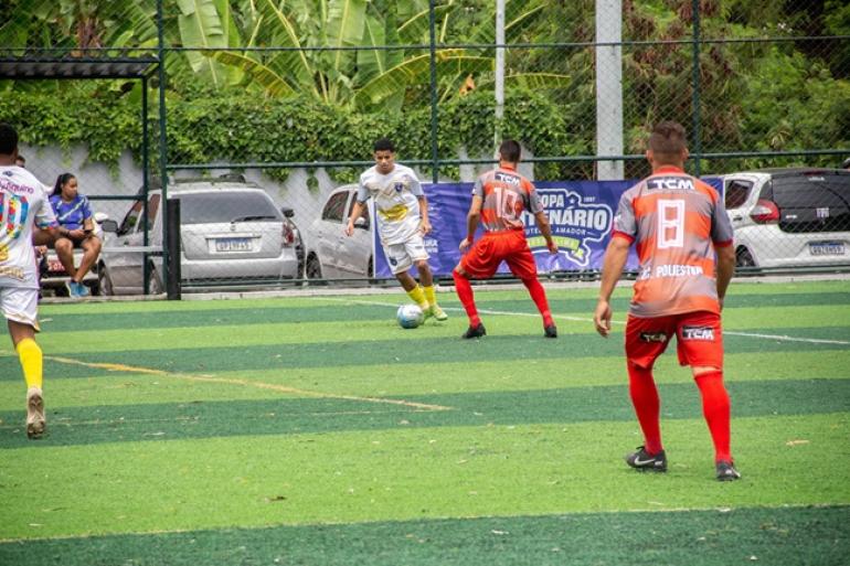 Em um campo de futebol com gramado sintético, jogadores disputam a final do Módulo C da Copa Centenário. Ao centro, um atleta de uniforme branco e amarelo conduz a bola enquanto é marcado por um jogador de uniforme cinza, vermelho e laranja. Outros dois atletas aparecem nas laterais da imagem. Ao fundo, atrás do alambrado, há árvores, carros estacionados e uma faixa da Copa Centenário de Futebol Amador Wadson Lima.