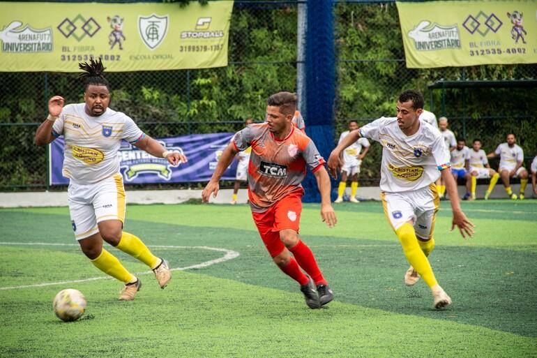 Em um campo de futebol society com gramado sintético, três jogadores disputam a bola. À esquerda, um atleta de uniforme branco, com detalhes dourados e meias amarelas, corre em direção à bola, que está à sua frente. No centro, um jogador com camisa cinza e laranja, calção vermelho e meias vermelhas acompanha a jogada. À direita, outro atleta de branco e amarelo também corre para alcançar a bola. Ao fundo, há jogadores reservas sentados no banco, alambrados, árvores e faixas de publicidade.