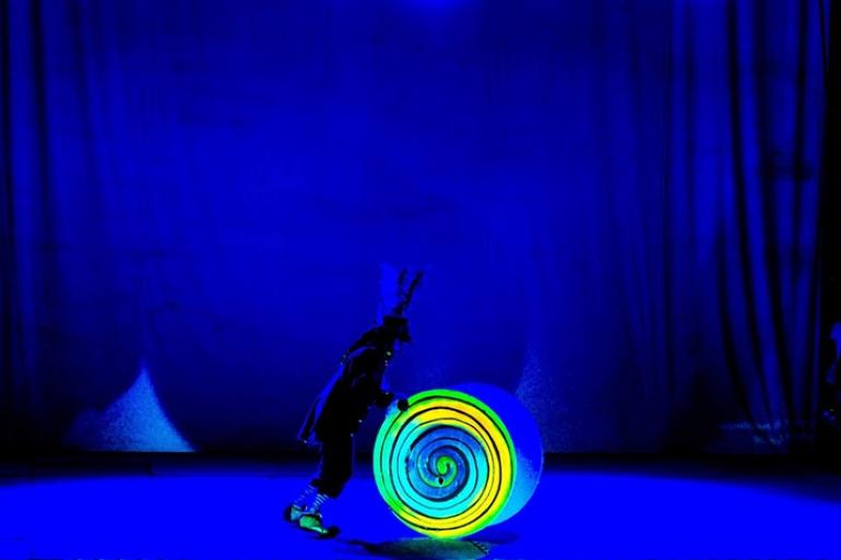 Em um palco iluminado por uma forte luz azul monocromática, um artista caracterizado empurra um grande cilindro decorado com uma espiral luminosa em tons de amarelo e verde. O performer veste casaco escuro, calças médias com meias listradas e um chapéu adornado com grandes penas. O fundo é composto por cortinas escuras sob a iluminação azul.