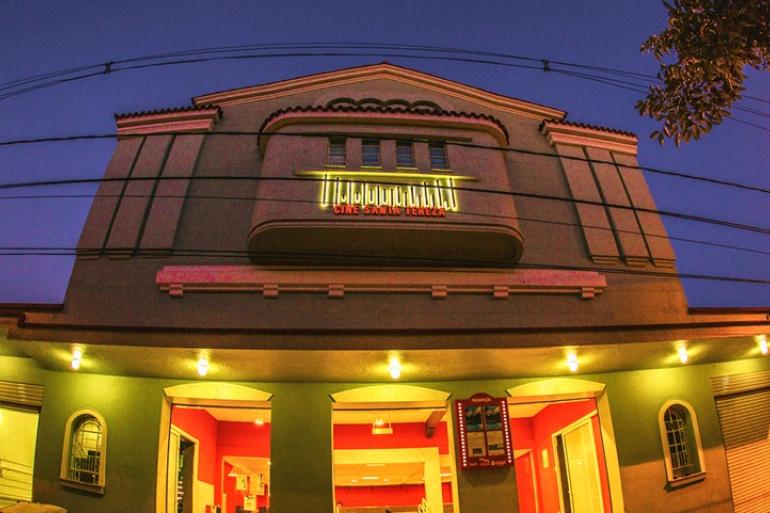 Fotografia da fachada do Cine Santa Tereza ao anoitecer, tirada de baixo para cima. O prédio tem um formato levemente curvado nas bordas por causa do efeito da foto. 
