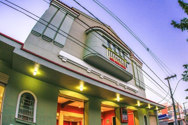 Fotografia em ângulo baixo da fachada do Cine Santa Tereza ao entardecer, prédio histórico em estilo Art Déco, em tom verde-claro, com frisos geométricos e um balcão curvado no centro, onde se destaca o letreiro vermelho e dourado com o nome do cinema.
