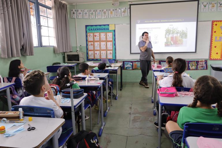 Fotografia de uma sala de aula, onde um professor está em pé à frente da turma, fazendo sinais em Libras. Ao redor, estudantes estão sentados em suas carteiras, acompanhando a aula. Ao fundo, há um projetor com uma apresentação intitulada “Aula de Libras – Ensino Fundamental 2”, além de cartazes educativos, calendário, livros e materiais escolares.