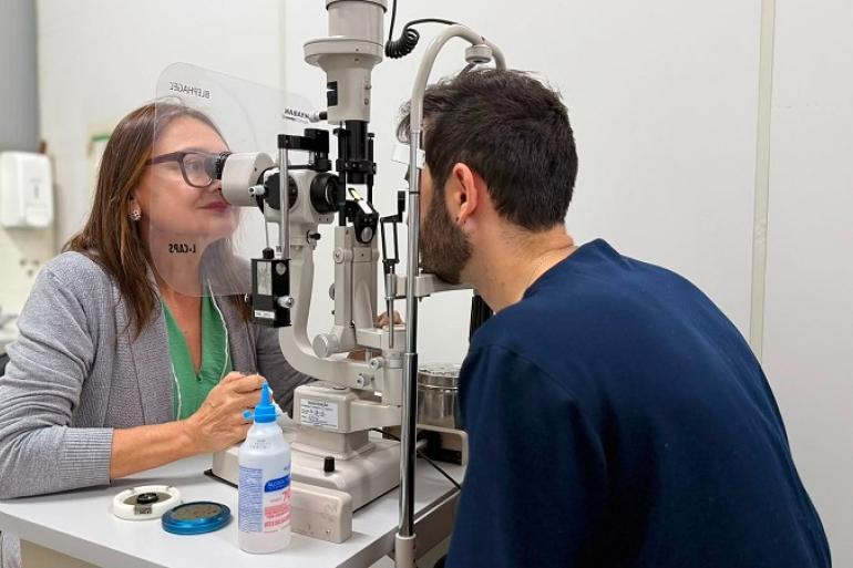 Médica oftalmologista vestida com blusa verde e com casaco cinza atende um paciente de barba vestido com uma blusa azul de mangas compridas.