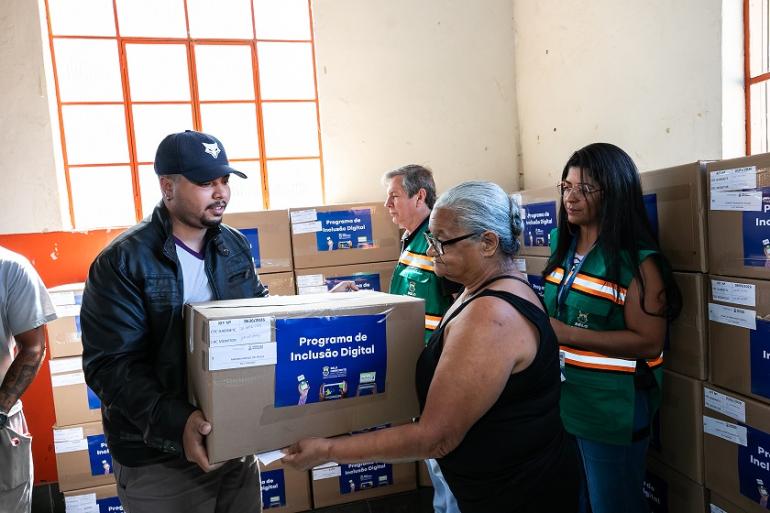 Mais cem equipamentos são entregues a famílias de Belo Horizonte pela Política de Inclusão Digital da Prefeitura Uma mulher de cabelos brancos recebe uma caixa de computador de um homem de casaco de couro, observados por um homem e uma mulher com colete da P)BH.