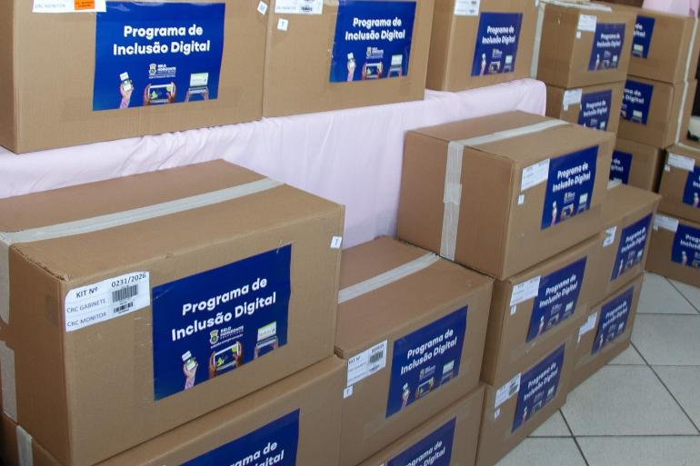 Caixas empilhadas com computadores na cor parda com adesivos em azul escrito Programa de Inclusão Digital