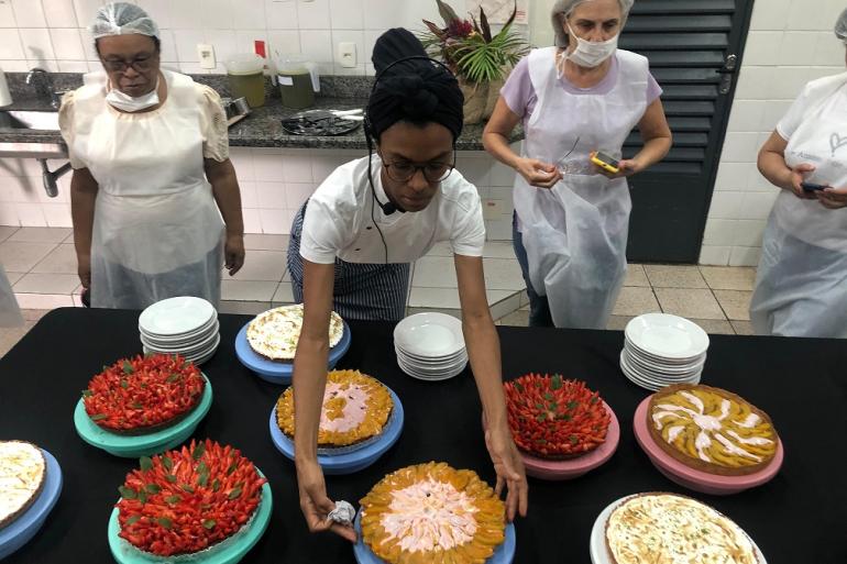 Imagem mostra quatro mulheres vestidas com dolmãs em frente ao mesa com várias tortas. Uma delas está manipulando uma das tortas enquanto as outras observam.