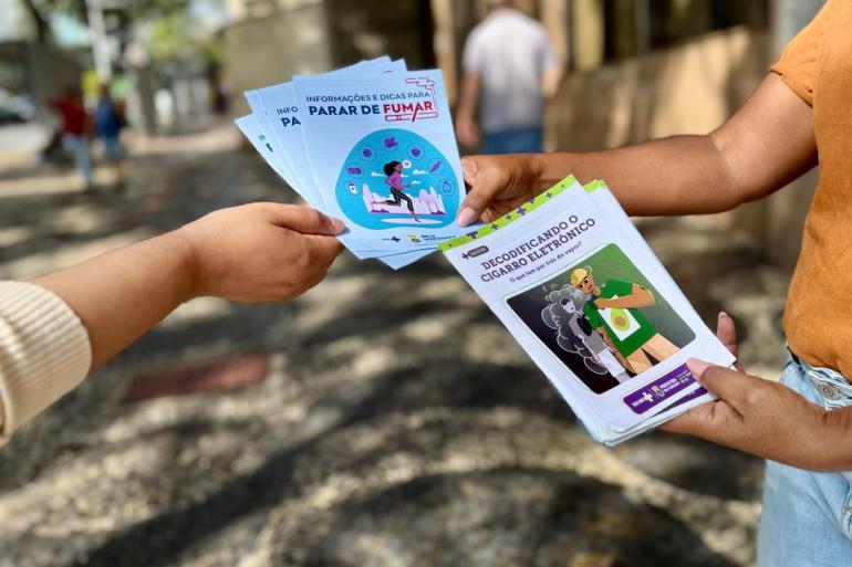 A foto mostra as mãos de duas pessoas segurando panfletos informativos, com orientações sobre os riscos do consumo do tabaco. 