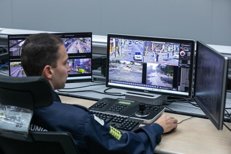 Muralha BH auxilia na localização de procurados pela Justiça e contribui para 22 prisões na capital Agente da Guarda Municipal, usando uniforme azul, está de frente para tela do computador fazendo o monitoramento das imagens geradas pelas câmeras do COP-BH.