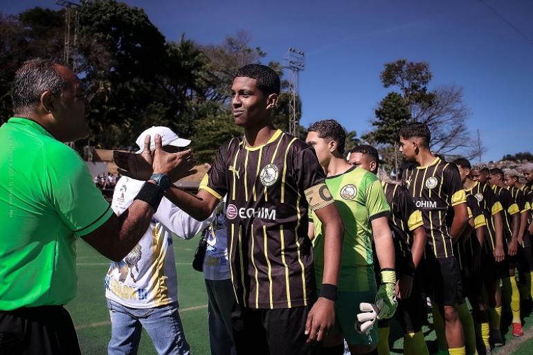 Copa Centenário terá finais do Sub-17 e do Master 40 neste sábado (8) Em um campo de futebol com gramado sintético, sob céu azul, um jogador jovem, de uniforme preto com listras verticais amarelas, cumprimenta o juiz da partida de camisa verde, que aparece de perfil à esquerda da imagem. O atleta está no centro, sorrindo discretamente, com a mão direita estendida durante o cumprimento. Atrás dele, jogadores da mesma equipe formam uma fila. A maioria usa camisa preta e amarela, calção preto e meiões amarelos. Logo ao lado do jogador central, o goleiro veste uniforme verde e lu
