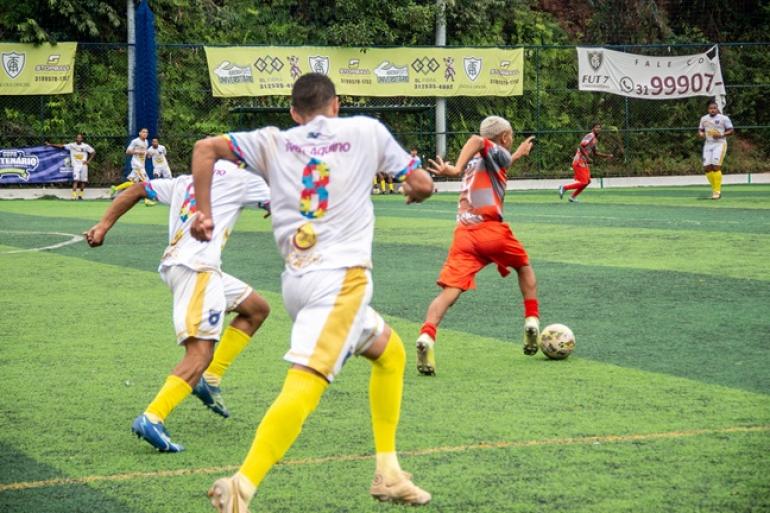 Copa Centenário terá 14 jogos decisivos neste domingo (27) nos módulos A, B e C Homens jogam futebol, um jogador de uniforme vermelho conduz a bola e é perseguido por dois homens de uniforme branco e amarelo