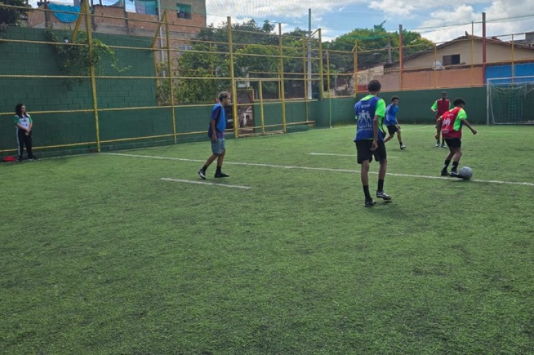 Copa do Mundo do Esporte Esperança movimenta a Arena Sumaré durante as férias escolares Jovens usando coletes azuis e vermelhos jogando futebol em um campo de gramado, enquanto professora observa a partida