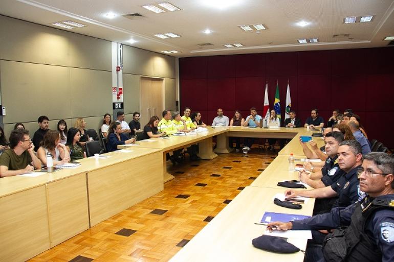 Imagem mostra várias pessoas participando de uma reunião em uma mesa retangular. Ao fundo há bandeiras , várias autoridades, entre elas, a subsecretária de Relações Internacionais da Prefeitura, Chyara Sales Pereira, responsável pela interlocução com a Fifa acerca da Copa do Mundo Feminina Fifa 2027.