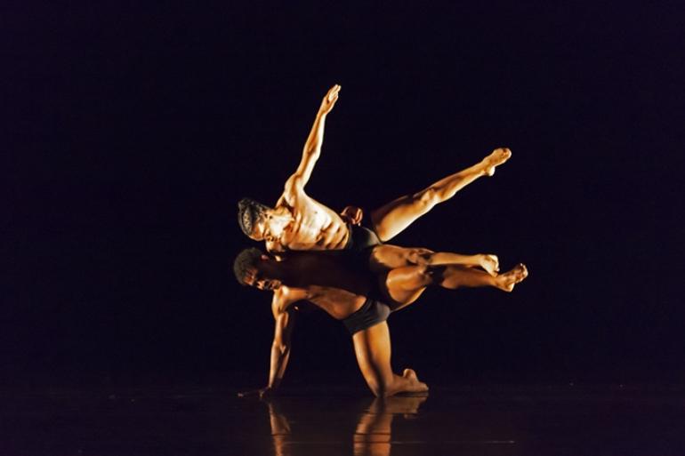 5ª Mostra CRDançaBH tem programação gratuita com espetáculos, residências, oficinas e debates Dois bailarinos aparecem em cena durante uma apresentação de dança contemporânea, iluminados por uma luz quente contra o fundo escuro do palco. Um bailarino está ajoelhado, com uma das mãos apoiada no chão, enquanto sustenta e se conecta ao corpo de outro bailarino de cabelos trançados. O segundo bailarino mantém o corpo inclinado horizontalmente