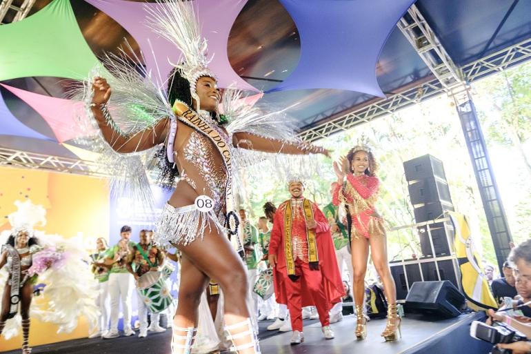 Rainha do Carnaval faz apresentação no palco durante o evento de eleição da Corte Momesca de 2026, sendo aplaudida pelos integrantes da Corte Momesca de 2025.