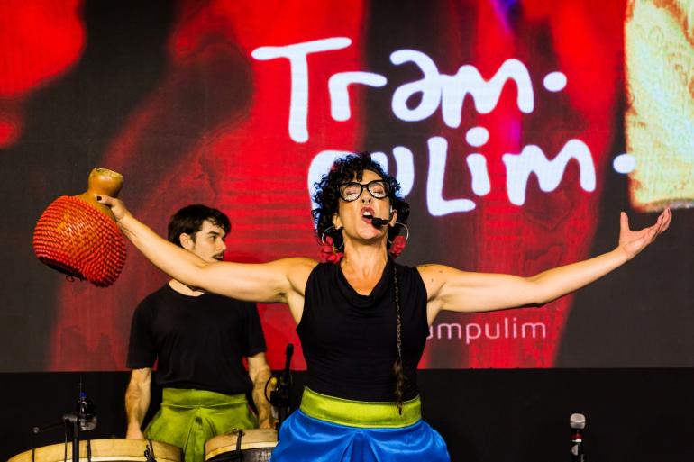 Uma artista no centro do palco expressa grande emoção ao cantar com a boca aberta e braços bem abertos. Ela tem cabelos cacheados pretos, usa óculos de grau, brincos grandes e blusa preta sem mangas. Em uma das mãos, segura um instrumento percussivo (xequerê) para o alto. Ao fundo, à esquerda, um homem de camiseta preta toca tambores. Atrás deles, um telão iluminado em tom vermelho exibe a palavra "Trampolim" escrita em letras brancas estilizadas.