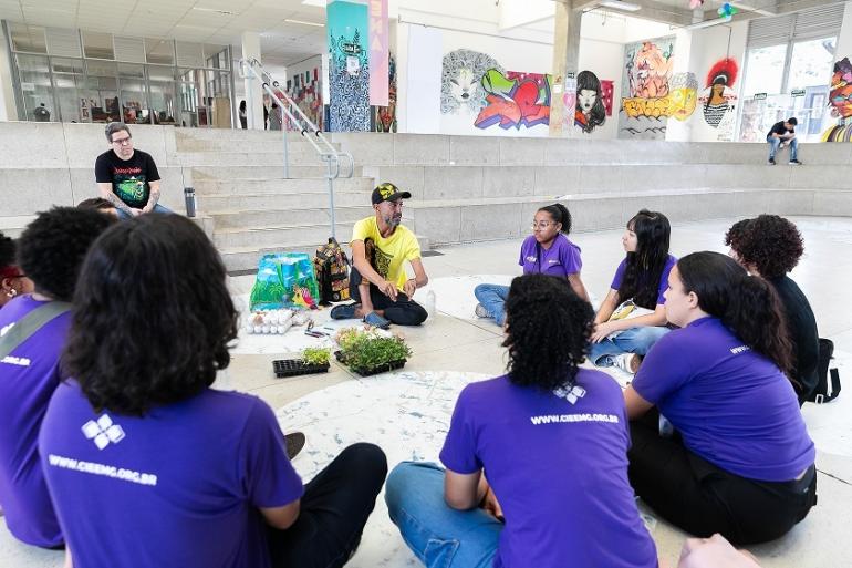 Semana das Juventudes terá Feirão de Empregos e debates sobre inteligência artificial Nove jovens e um educador posam agachados em círculo, segurando pequenas mudas de plantas. No centro, há bandejas com mudas, cartelas de ovos e materiais utilizados na atividade. Ao fundo, o espaço do CRJ exibe murais coloridos e um auditório.