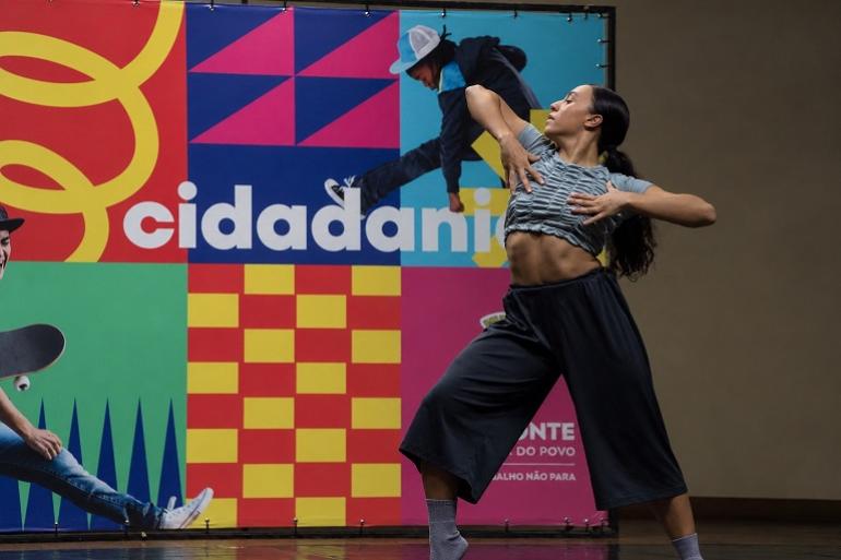 Aula aberta de dança é destaque da programação desta semana no Centro de Referência das Juventudes Em um espaço interno, uma mulher dança em primeiro plano, com o corpo inclinado para trás e os braços erguidos junto ao peito. Ela veste blusa cinza franzida, calça preta ampla e meias cinzas. Ao fundo, há um painel grande e colorido, formado por blocos em vermelho, azul, amarelo, verde e rosa, com a palavra “cidadania” em destaque. O painel também traz imagens de pessoas em movimento. A iluminação é baixa, com maior destaque sobre a dançarina.