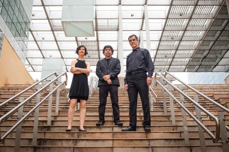 A foto mostra uma mulher e dois homens, todos vestidos com trajes pretos, posando em pé sobre os degraus de uma larga escadaria ao ar livre. À esquerda da imagem, a mulher usa vestido preto sem mangas e tem os braços cruzados; no centro, há um homem de terno preto com as mãos entrelaçadas à frente; à direita, o outro homem está de camisa de manga longa e calça pretas. Ao fundo, destaca-se uma estrutura arquitetônica moderna com teto de vidro e suportes metálicos que cobrem o espaço.